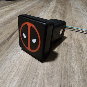 May include: Black trailer hitch cover with a red and white Deadpool logo.