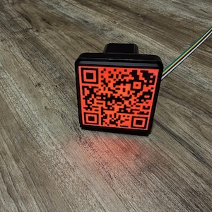 May include: A black square-shaped device with a red illuminated QR code. The device is mounted on a wooden surface.