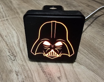 Puede incluir: Una cubierta negra para enganche de remolque con el contorno naranja brillante del casco de Darth Vader. El diseño está iluminado con luz naranja. La cubierta es rectangular con un poste de montaje.