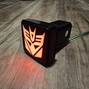 Può includere: Copertura nera per gancio di traino con un simbolo Autobot arancione brillante. La copertura quadrata ha una cornice nera e un emblema illuminato. I fili si estendono dalla parte posteriore. L'articolo è su una superficie di legno.