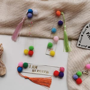 May include: A set of six colourful bookmarks with pom-poms. The bookmarks are made of clear acrylic and feature different inspirational messages, including "I am Blessed" and "Faith".