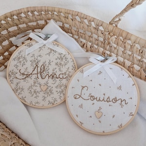 Peut inclure: Deux cercles brodés avec les noms "Alma" et "Louison" en écriture cursive. Les cercles ont un tissu floral, des cadres en bois et des rubans blancs. Un petit cœur en bois est sous chaque nom.