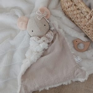 Peut inclure: Une couverture de sécurité en peluche avec une tête de souris beige aux oreilles roses, un nœud floral et un col à volants. La couverture présente un tissu beige texturé à petits pois d'un côté et de la fausse fourrure blanche, avec l'étiquette Zoli & Compagnie.