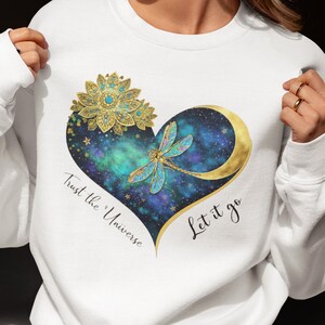 May include: A white long-sleeve sweatshirt featuring a central heart-shaped graphic. The heart contains a blue and green nebula pattern, a gold dragonfly, a gold crescent moon, and a gold lotus flower. The text reads Trust the Universe Let it go.