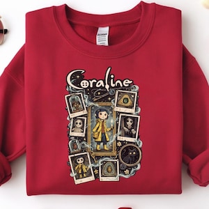 Op de afbeelding: Een donkerrood sweatshirt met ronde hals en een grafische print van het personage Coraline. Het ontwerp bevat de tekst Coraline, een centrale illustratie van het personage in een gele regenjas en diverse ingelijste afbeeldingen van figuren.