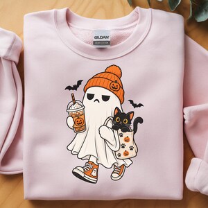 May include: A light pink crewneck sweatshirt featuring a graphic of a white ghost wearing an orange beanie and orange sneakers. The ghost holds a coffee cup with a pumpkin design and carries a tote bag containing a black cat with pumpkin prints.