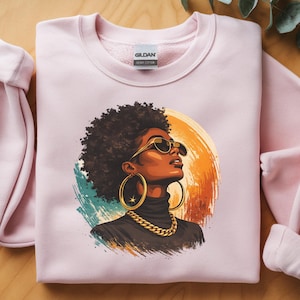 May include: A light pink crewneck sweatshirt featuring a graphic illustration of a person with dark curly hair, wearing large gold hoop earrings, a thick gold chain necklace, and dark sunglasses, set against a circular orange and teal background.