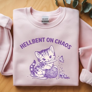 May include: A light pink sweatshirt with the text "HELLBENT ON CHAOS" above a purple illustration of a kitten holding a knife and a ball of yarn. A butterfly and sewing notions are also in the graphic.