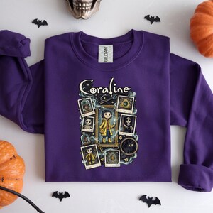 Op de afbeelding: Een paars sweatshirt met lange mouwen en een grafische print van het personage Coraline. Het ontwerp bevat de tekst Coraline, een centrale illustratie van het personage in een gele regenjas en diverse ingelijste afbeeldingen van figuren en objecten.
