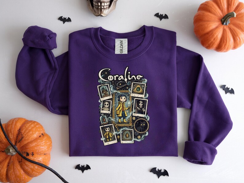 Op de afbeelding: Een paars sweatshirt met lange mouwen en een grafische print van het personage Coraline. Het ontwerp bevat de tekst Coraline, een centrale illustratie van het personage in een gele regenjas en diverse ingelijste afbeeldingen van figuren en objecten.