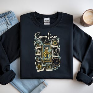 Op de afbeelding: Een zwart sweatshirt met ronde hals en een grafische print met de tekst Coraline. Het ontwerp bevat een centrale illustratie van een personage in een gele regenjas, omringd door verschillende ingelijste polaroid-stijl afbeeldingen van figuren.