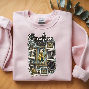 Op de afbeelding: Een lichtroze sweater met ronde hals en een grafische print van het personage Coraline. Het ontwerp bevat de tekst Coraline, een centrale illustratie van het personage in een gele regenjas en verschillende ingelijste afbeeldingen van figuren.