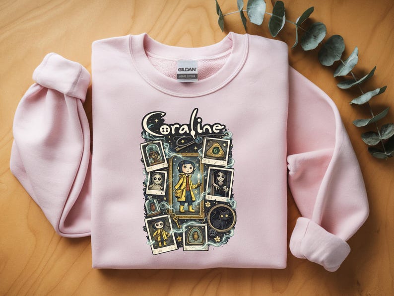 Op de afbeelding: Een lichtroze sweater met ronde hals en een grafische print van het personage Coraline. Het ontwerp bevat de tekst Coraline, een centrale illustratie van het personage in een gele regenjas en verschillende ingelijste afbeeldingen van figuren.
