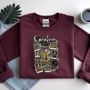 Op de afbeelding: Een bordeauxrood sweatshirt met lange mouwen en een grafische print van het personage Coraline. Het ontwerp bevat de tekst Coraline, een centrale illustratie van het personage in een gele regenjas en diverse ingelijste afbeeldingen.