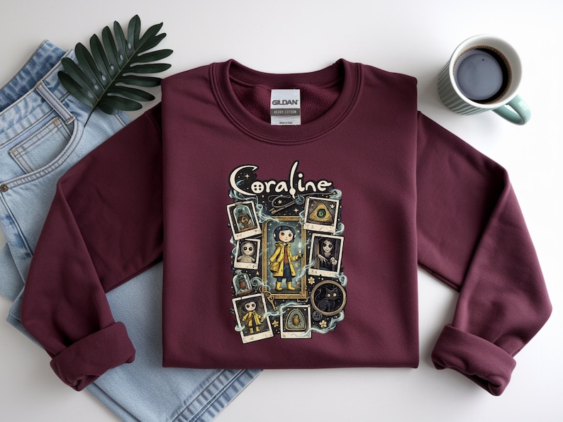 Op de afbeelding: Een bordeauxrood sweatshirt met lange mouwen en een grafische print van het personage Coraline. Het ontwerp bevat de tekst Coraline, een centrale illustratie van het personage in een gele regenjas en diverse ingelijste afbeeldingen.