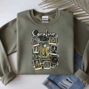 Op de afbeelding: Een olijfgroene sweater met ronde hals en een grafische print met de tekst Coraline. Het ontwerp bevat een centrale illustratie van een personage in een gele regenjas, omringd door diverse ingelijste afbeeldingen, waaronder een zwarte kat en knopen.