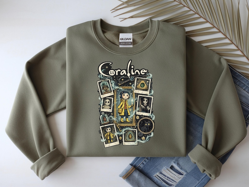 Op de afbeelding: Een olijfgroene sweater met ronde hals en een grafische print met de tekst Coraline. Het ontwerp bevat een centrale illustratie van een personage in een gele regenjas, omringd door diverse ingelijste afbeeldingen, waaronder een zwarte kat en knopen.