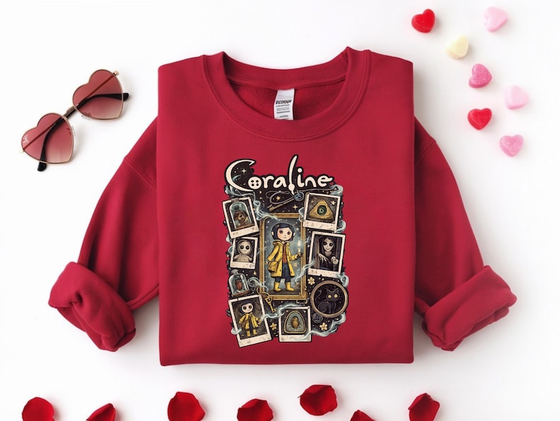 Op de afbeelding: Een donkerrood sweatshirt met ronde hals en een grafische print van het personage Coraline. Het ontwerp bevat de tekst Coraline, een centrale illustratie van het personage in een gele regenjas en diverse ingelijste afbeeldingen van figuren.