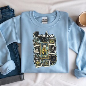 Op de afbeelding: Een lichtblauw sweatshirt met ronde hals en een grafische print van het personage Coraline. Het ontwerp bevat de tekst Coraline, een centrale illustratie van het personage in een gele regenjas en diverse ingelijste afbeeldingen van personages en objecten.