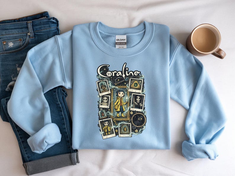 Op de afbeelding: Een lichtblauw sweatshirt met ronde hals en een grafische print van het personage Coraline. Het ontwerp bevat de tekst Coraline, een centrale illustratie van het personage in een gele regenjas en diverse ingelijste afbeeldingen van personages en objecten.