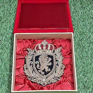 Peut inclure: Un écusson brodé décoratif représentant un lion doré sur un bouclier noir, surmonté d'une couronne et entouré de filigrane doré. L'écusson est présenté dans une boîte carrée avec une doublure en velours rouge et un couvercle rouge articulé.