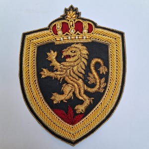 May include: Embroidered patch featuring a golden lion rampant on a black shield, topped with a crown. The shield is outlined with gold thread and a black border. Red accents are visible on the lion's tongue and at the base of the shield.