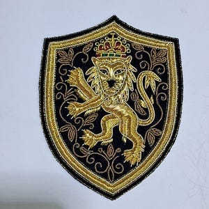 May include: Embroidered patch featuring a golden lion wearing a crown, set against a black shield-shaped background. The lion is intricately detailed with golden thread, surrounded by gold trim and decorative floral elements. The patch is designed for application on clothing or accessories.