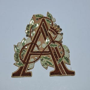 May include: Embroidered letter "A" in brown, adorned with green leaves, sequins, and beads. The letter has a light brown background and is embellished with intricate details, creating a decorative and elegant design.