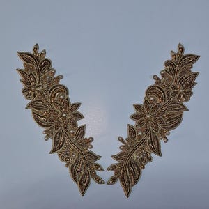 May include: Two ornate, symmetrical appliques with a floral and leaf design. The appliques are primarily gold and brown, with small pearl-like beads. The design features detailed embroidery and a delicate, textured appearance.