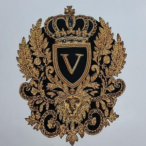 May include: An ornate, embroidered emblem featuring a black shield with a gold "V" and a gold crown. The emblem is surrounded by gold leaves, a lion's head, and decorative scrollwork. The design is rich in detail and texture.