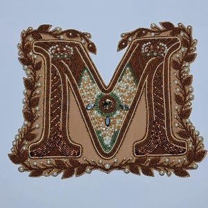 Puede incluir: Letra "M" bordada en tonos marrón, dorado y crema. El diseño incorpora perlas, cuentas doradas y una corona. Un borde de hojas enmarca la letra, creando un monograma decorativo.
