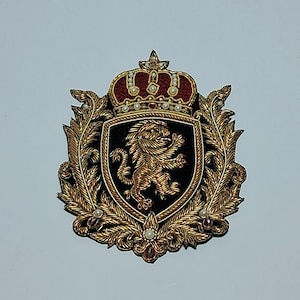 May include: An ornate, embroidered crest featuring a gold lion rampant on a black shield, topped with a red and gold crown. The crest is surrounded by gold leaves and embellished with pearls and red jewels, creating a regal design.