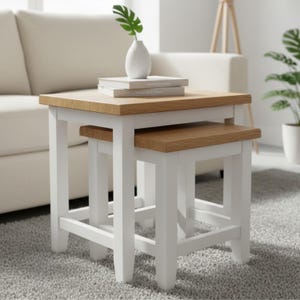 May include: Two square nesting tables, white-painted base with natural wood tops. The larger table is about 61 cm wide and 51 cm tall. A small white vase with a green plant and books are on the top table. The tables are on a grey carpet.