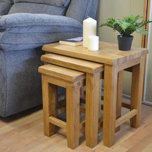 May include: Three wooden nesting tables in a natural finish. The tables are rectangular with solid legs. A small potted plant, two white candles, and a book are on the largest table. The tables are near a grey sofa.