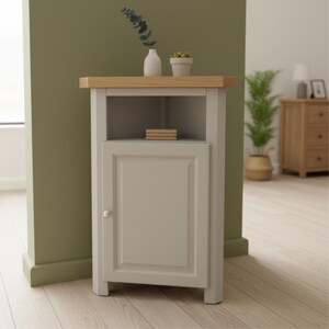 May include: A light grey corner cabinet featuring a natural wood top, an open upper shelf, and a single lower cabinet door with a small round knob. The cabinet has a panelled door design and stands on four legs, providing storage space for home items.