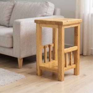 May include: A light-coloured wooden side table with a square top and a lower shelf. The table has a simple, sturdy design with vertical supports and a natural wood grain finish. The table is next to a light-coloured sofa.