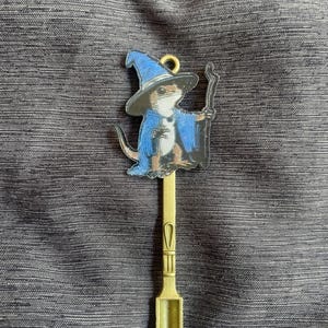 May include: A decorative spoon featuring a wizard lizard. The lizard is dressed in a blue wizard hat and robe, holding a staff. The spoon is gold-coloured and has a scoop at the end.