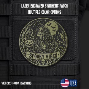 May include: A circular olive green laser engraved synthetic patch with a hook backing. The design features a skeleton in a hooded cloak holding a jack-o'-lantern and a candle, with a haunted house, bats, and the text SPOOKY VIBES printed below.