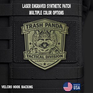 May include: An olive green shield-shaped patch with a laser-engraved raccoon wearing a tactical helmet. The text reads TRASH PANDA TACTICAL DIVISION ALWAYS LOOTING. The patch features a hook backing and is displayed on a black tactical gear surface.