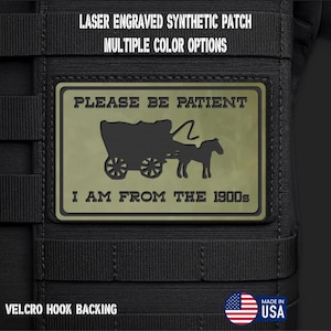 May include: A rectangular olive green synthetic patch with a black laser-engraved graphic of a horse-drawn carriage. The text reads PLEASE BE PATIENT I AM FROM THE 1900s. The patch features a hook backing and is attached to a black tactical gear surface.