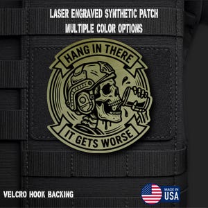May include: A circular olive green laser engraved synthetic patch featuring a skull wearing a tactical helmet and drinking from a cup. The text reads HANG IN THERE IT GETS WORSE. The patch is attached to a black fabric surface with velcro hook backing.