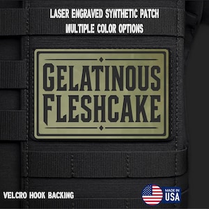 May include: A rectangular olive green laser engraved synthetic patch with black text reading GELATINOUS FLESHCAKE. The patch is attached to a black tactical gear surface with horizontal webbing. Text above reads LASER ENGRAVED SYNTHETIC PATCH MULTIPLE COLOR OPTIONS and below reads VELCRO HOOK BACKING with a USA flag icon.