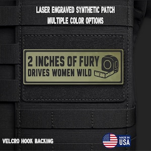 May include: A rectangular olive green laser engraved synthetic patch with a black border and black text that reads 2 INCHES OF FURY DRIVES WOMEN WILD, featuring a graphic of a measuring tape. The patch has a hook backing and is attached to black fabric.