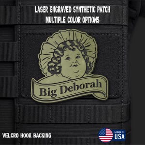 May include: A laser engraved synthetic patch featuring a stylized illustration of a person with curly hair and a bonnet, with a banner below reading Big Deborah. The patch is olive green with black details and includes a velcro hook backing.