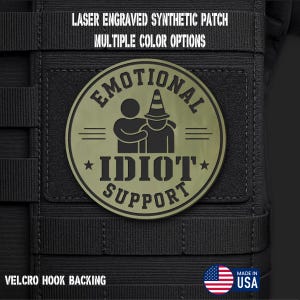 May include: A circular olive green laser engraved synthetic patch with a Velcro hook backing. The patch features a graphic of one figure comforting another wearing a cone hat, with the text "EMOTIONAL IDIOT SUPPORT" and stars.