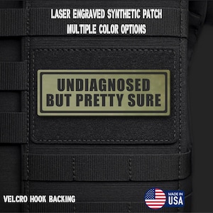 May include: A rectangular olive green synthetic patch with a black border and the text "UNDIAGNOSED BUT PRETTY SURE" in bold black letters. The patch features a hook backing and is attached to a black tactical gear surface with webbing straps.
