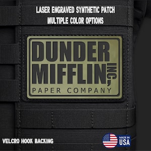 May include: A rectangular olive green synthetic patch with black laser-engraved text reading DUNDER MIFFLIN PAPER COMPANY. The patch is attached to a black tactical gear surface with horizontal webbing straps and features a velcro hook backing.