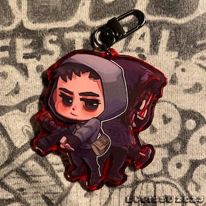 Könnte beinhalten: Ein rot umrandeter Acryl-Anhänger mit einer Cartoon-Figur in einer Kapuzenjacke, die eine Waffe hält. Der Anhänger hat einen schwarzen Verschluss und eine kleine, beige Umhängetasche. Die Figur befindet sich in Kampfhaltung.
