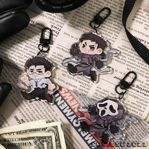 May include: Clear acrylic keychains featuring cartoon characters with black metal clasps. One keychain depicts a character with glasses, another with a peace sign, and a third with a knife. The keychains are on a newspaper background.
