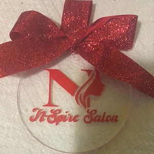 May include: A red glitter bow is tied around a clear round ornament with a red glitter background. The ornament has a red silhouette of a woman's head with a stylized letter N and the text "N-Spire Salon".